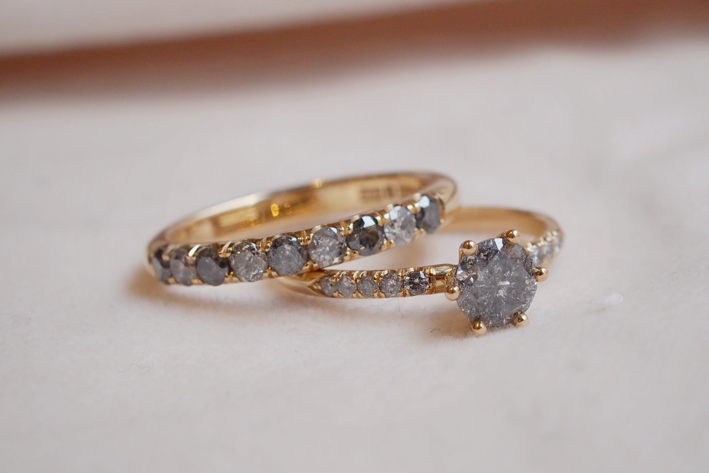 En gnistrande duo av diamantringar i 18k guld perfekta att kombinera med varandra. Den ena med en 0,70ct briljantslipad grå salt- & peppardiamant infattad i en sexklofattning, inramad av fem gråa briljantslipade diamanter i castellfattning på vardera sida. Den andra en alliansring med nio gråa diamanter i växlande ljusare och mörkare grå nyans, infattade i vacker kastellfattning. Närbild av vigselring och förlovningsring designad och tillverkade på Södermalm i Stockholm..
