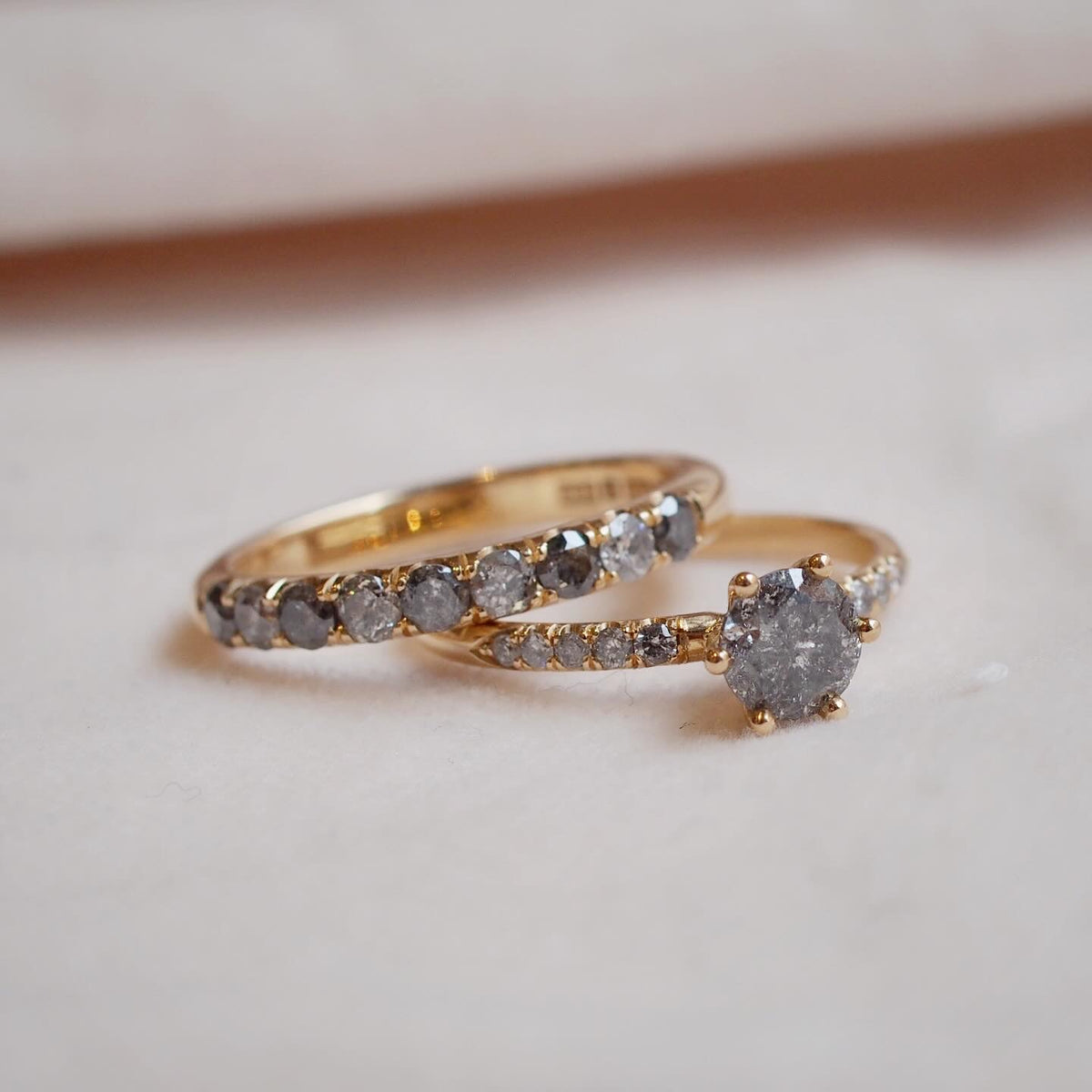 En duo av diamantringar i 18k guld perfekta att kombinera med varandra. Vigselring, förlovningsring eller en vardagslyxig present till dig själv. Den ena med en 0,70ct briljantslipad grå salt- & peppardiamant infattad i en sexklofattning, inramad av fem gråa briljantslipade diamanter i castellfattning på vardera sida. Den andra en alliansring med nio gråa diamanter i växlande ljusare och mörkare grå nyans, infattade i vacker kastellfattning. Närbild av förlovnings- och vigselringar.